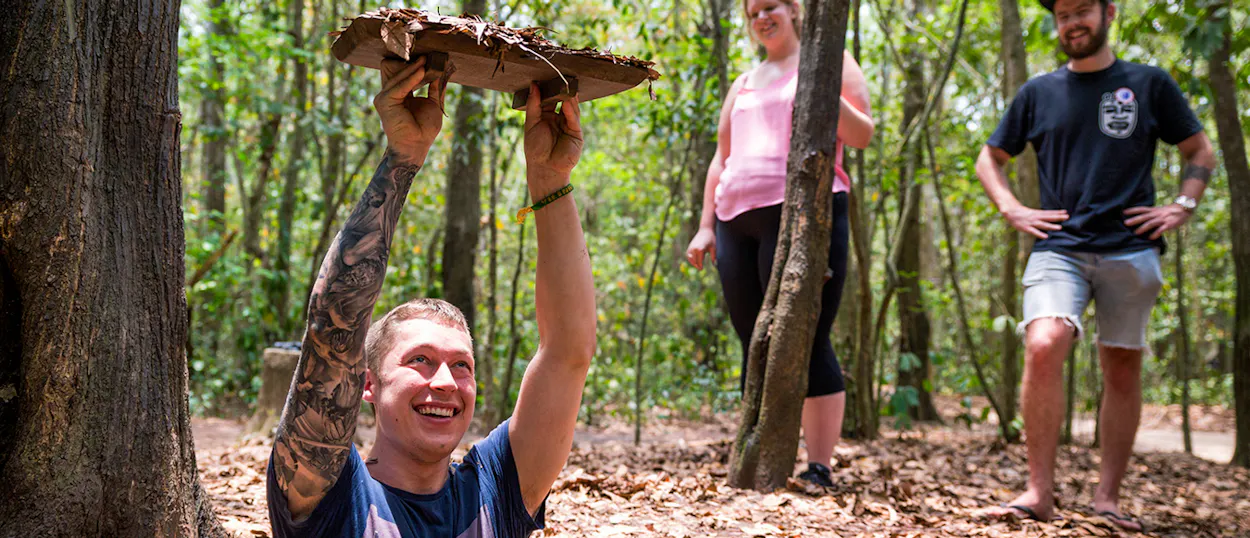 A smiling man with tattooed arms emerges from a camouflaged Cu Chi Tunnel entrance in a lush jungle.