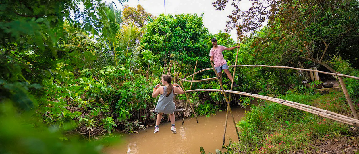 Two backpackers carefully cross a rustic bamboo bridge over a muddy canal in the Mekong Delta.