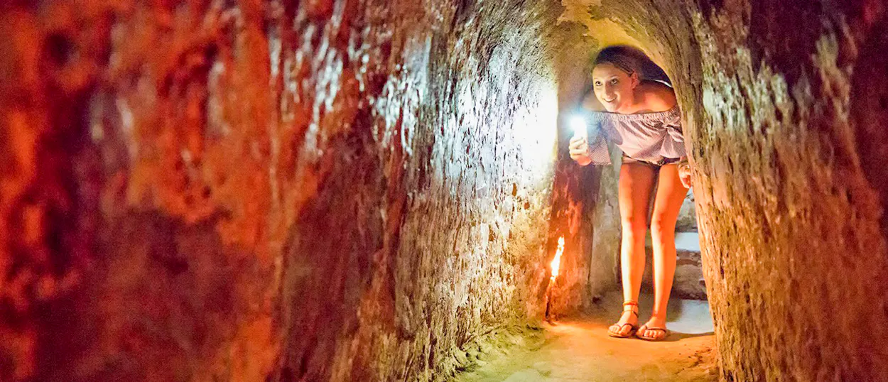 A young woman explores the narrow Cu Chi Tunnels in Vietnam, illuminating her path with a flashlight.