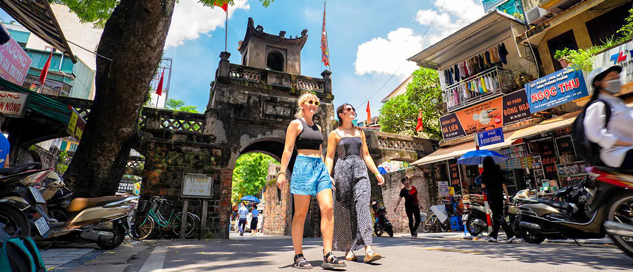 Two happy backpackers walk past an ancient city gate on a sunny street in Hanoi.