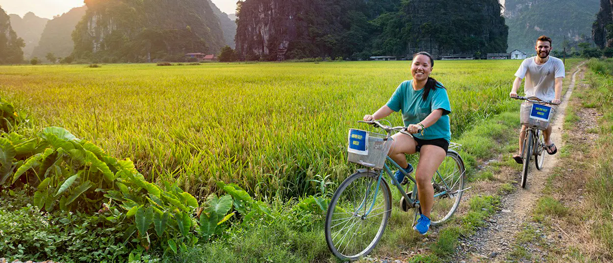 Two happy cyclists ride bikes through vibrant green rice paddies with majestic mountains in Ninh Binh.