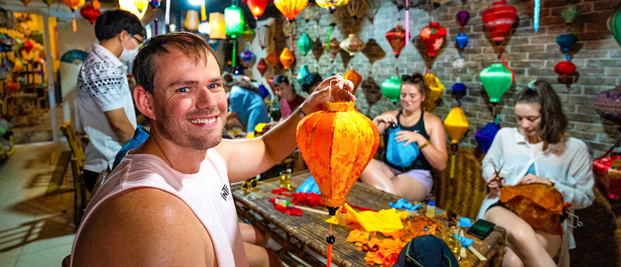 A smiling man proudly displays his handmade orange lantern in a vibrant Hoi An craft workshop.