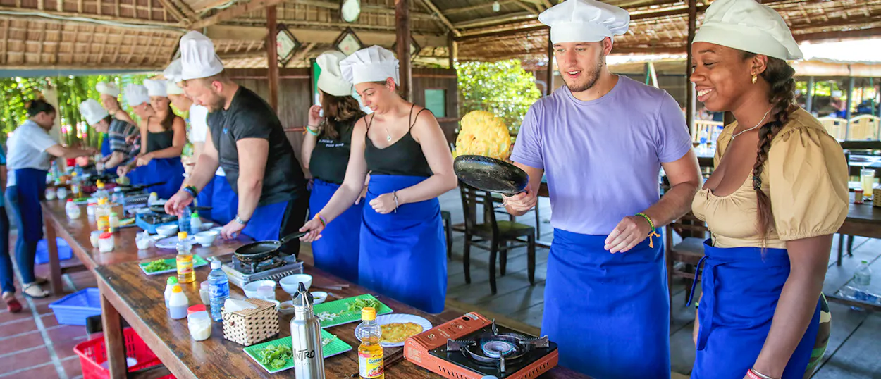 Smiling travelers in chef hats and aprons learning to cook at an outdoor class in Hoi An.