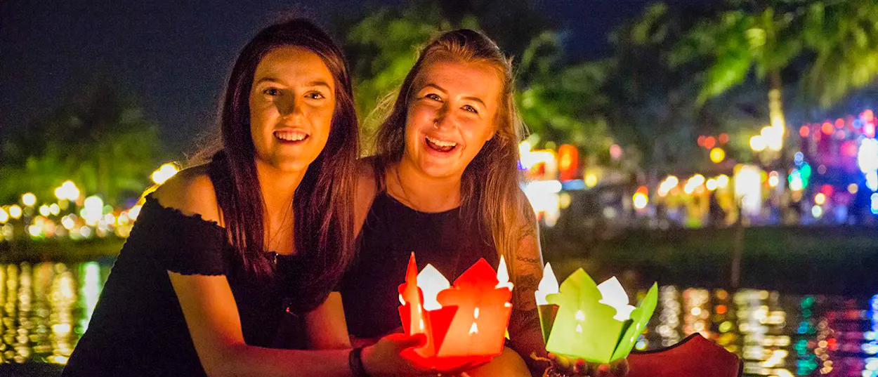 Two happy women holding glowing lanterns on a river in Hoi An at night.