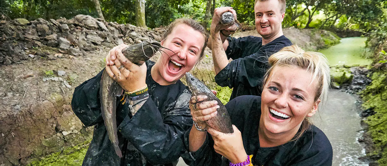 Mud-covered travelers laugh excitedly while holding their freshly caught fish in the Mekong Delta.