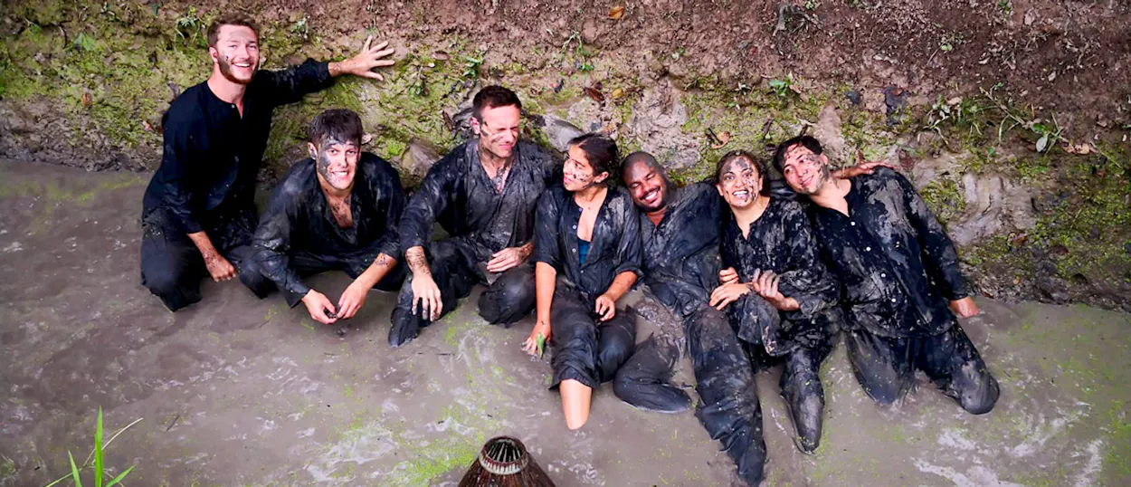 A group of happy travelers covered in mud, smiling while sitting in a muddy stream in the Mekong Delta.