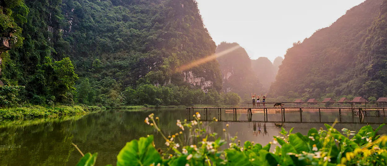 People walk on a wooden bridge over a river at sunset, surrounded by dramatic limestone mountains in Ninh Binh.