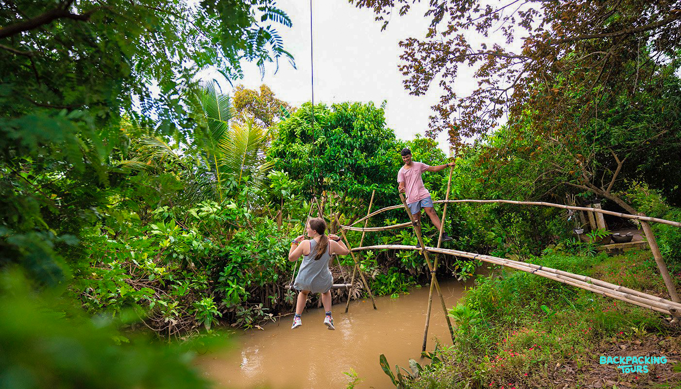Adventurous travelers cross a narrow bamboo bridge and swing over a muddy river in the Mekong Delta.