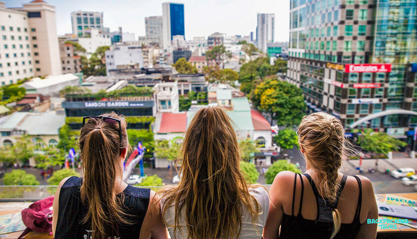 Three women enjoy a panoramic city view from a rooftop during their backpacking adventure.