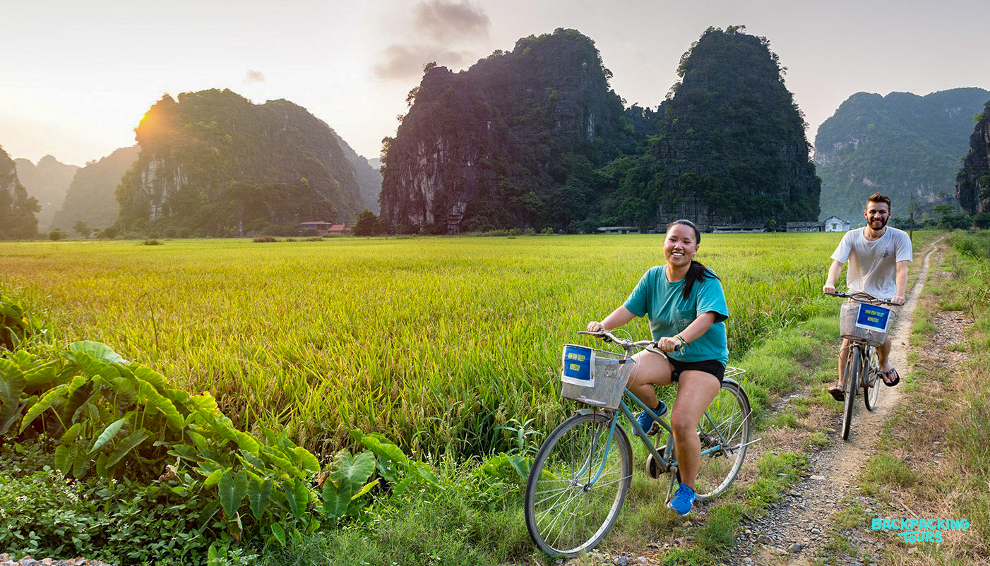 Two smiling travelers cycle through vibrant rice paddies with towering limestone karsts in Ninh Binh.