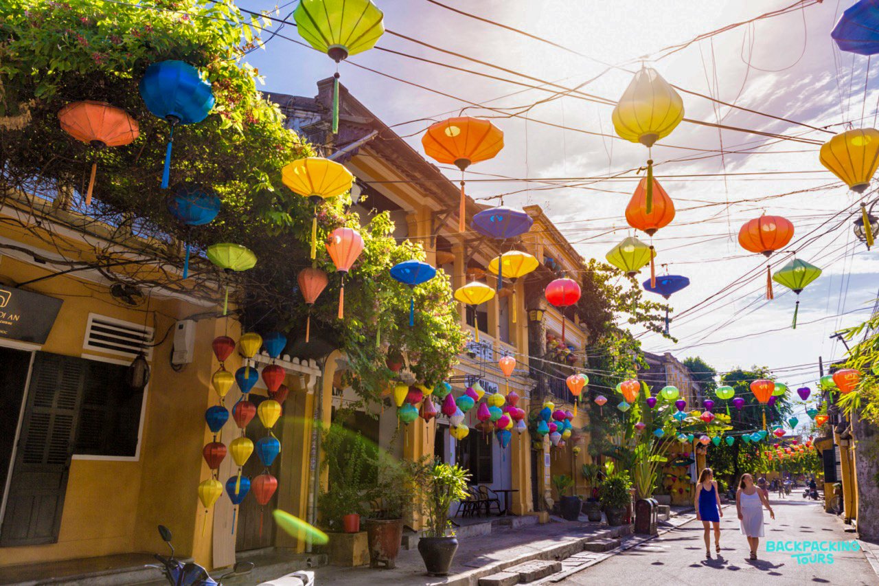 Colorful lanterns illuminate a charming street in Hoi An, with two people strolling in the afternoon sun.