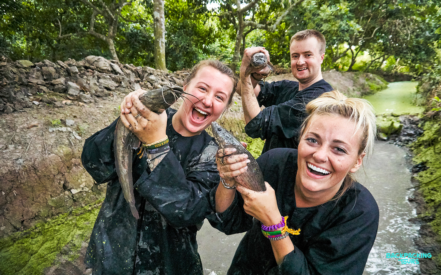 Three smiling backpackers covered in mud, proudly holding their freshly caught fish in the Mekong Delta.