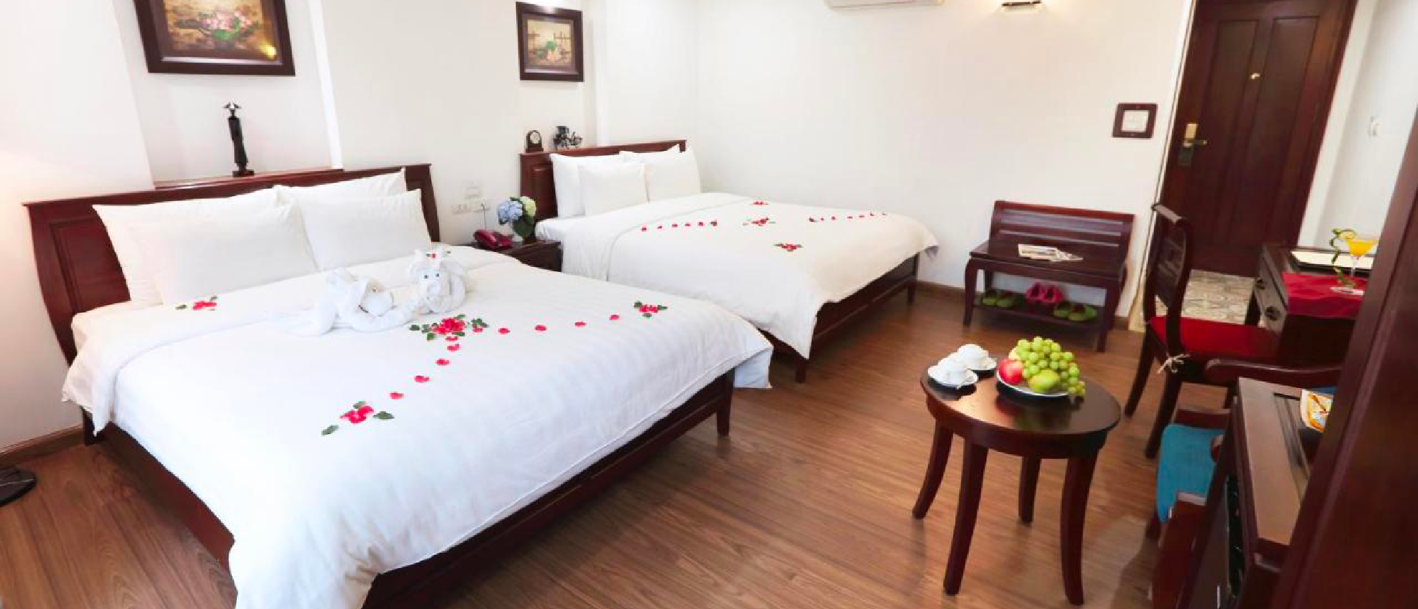 A clean, welcoming hotel room with two beds decorated with flower petals and a fruit bowl.