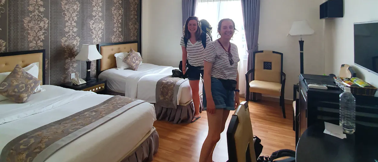 Two smiling women stand in a comfortable hotel room with two beds in Hue, Vietnam.