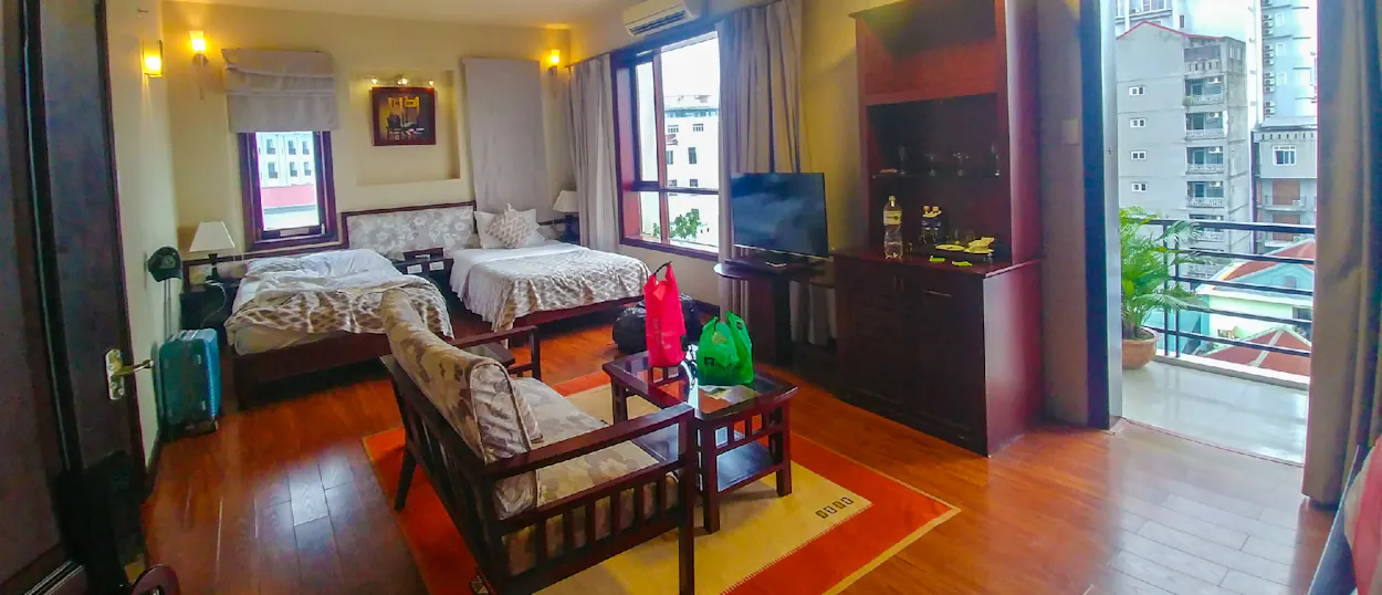 A spacious hotel room with two beds, a sofa, a TV, and a balcony overlooking city buildings.