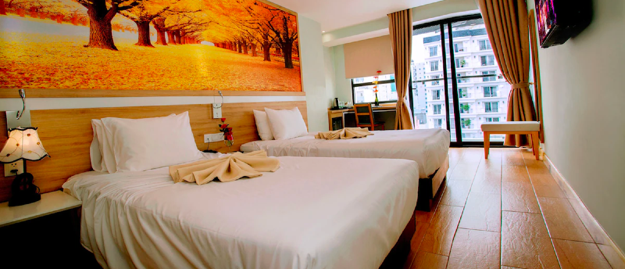 A bright hotel room in Nha Trang with two single beds, a colorful tree mural, and a city view.