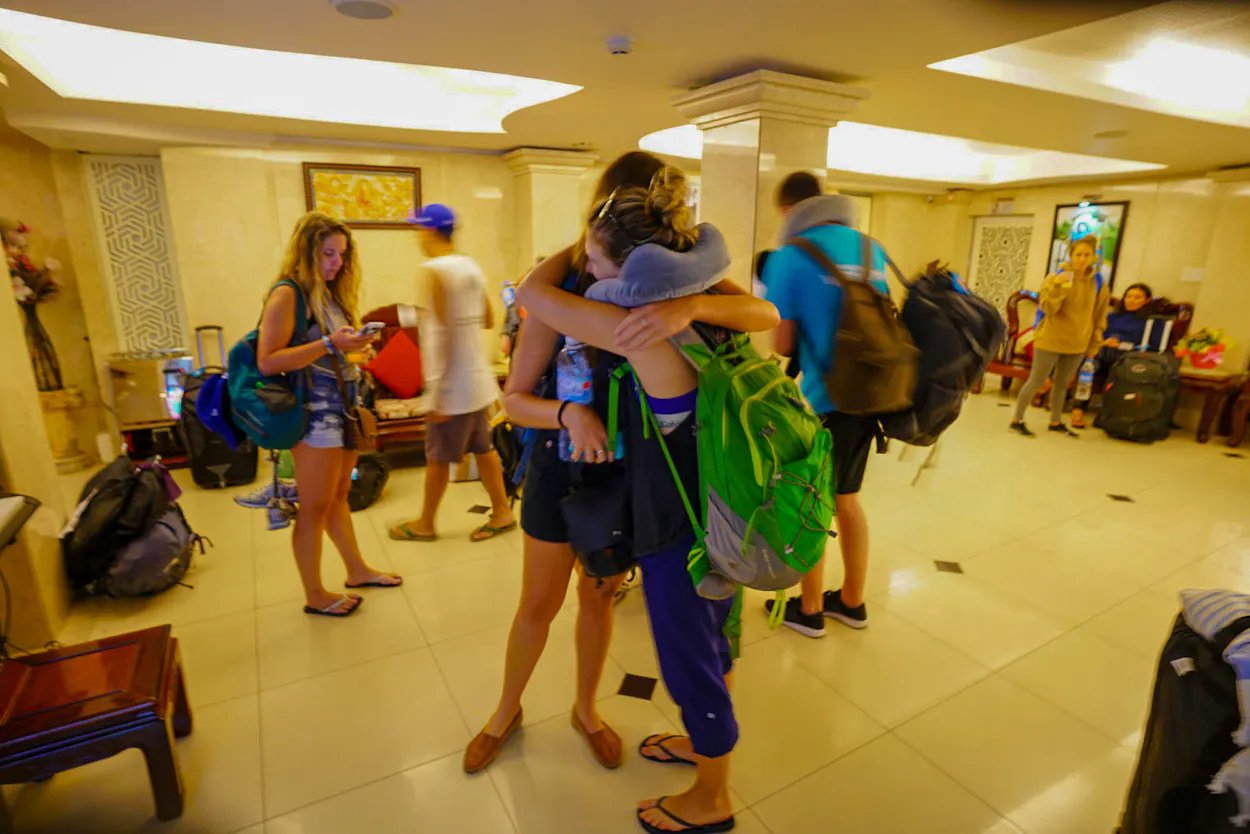 Backpackers share an emotional goodbye hug in a brightly lit hotel lobby with their gear.