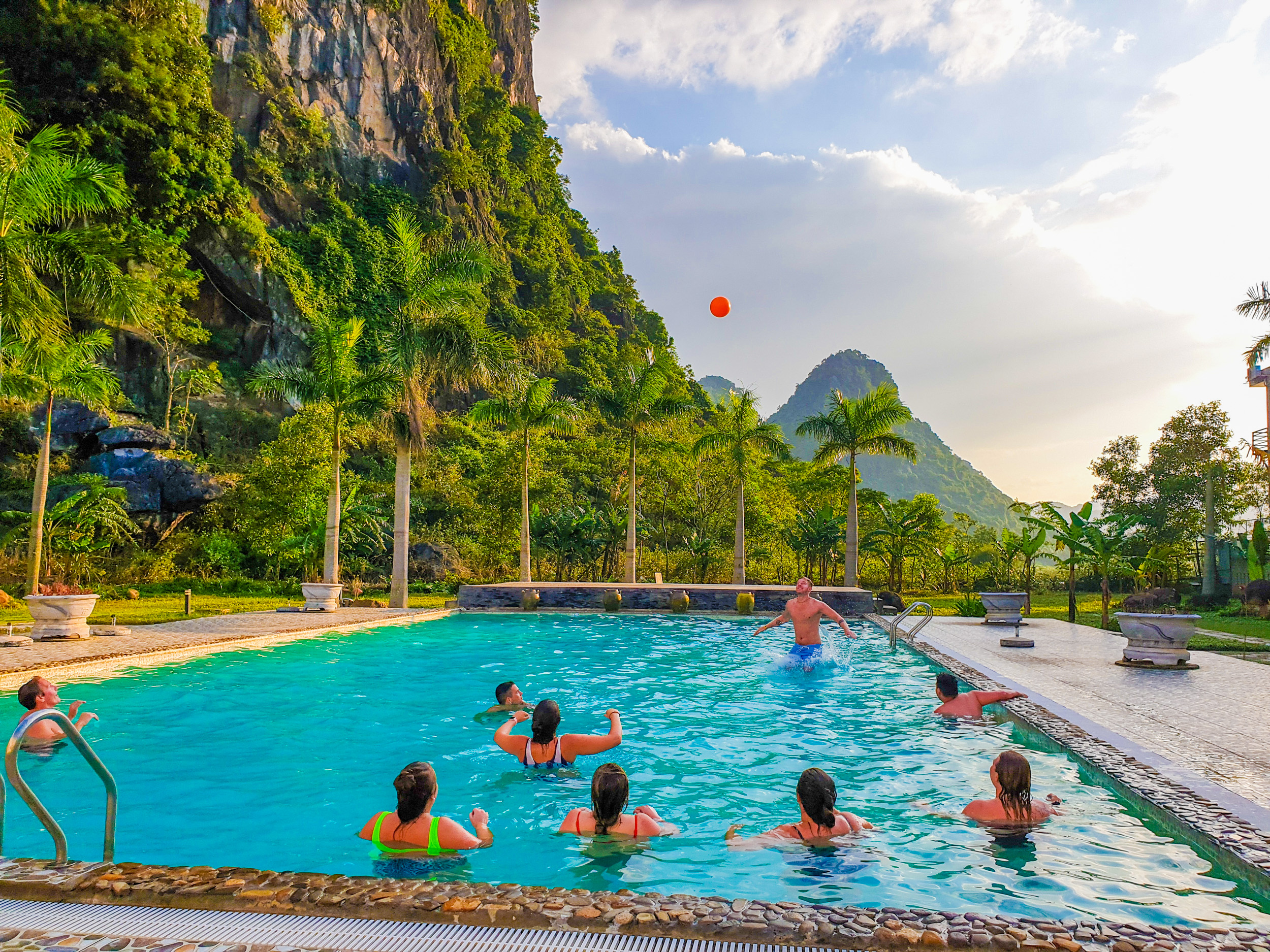 Travelers enjoy a game with an orange ball in a swimming pool amidst green mountains in Vietnam.